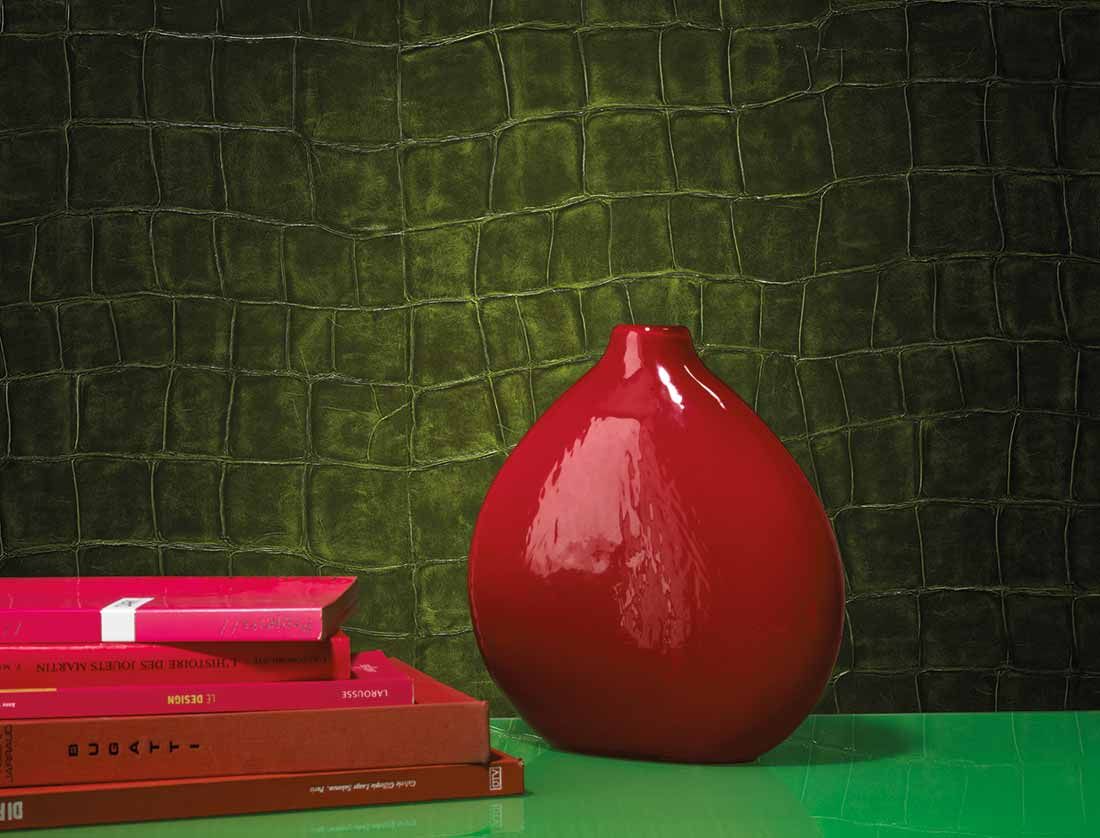 Eine grüne Krokodilhaut-Tapete in Lederoptik, davor eine rote Vase und rote Bücher