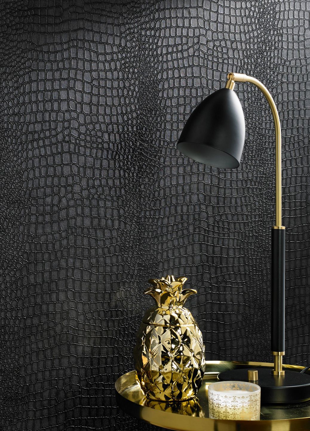 Papel de parede com aparência de couro negro de crocodilo por trás de uma mesa de apoio dourada com um candeeiro de mesa e um ananás decorativo dourado