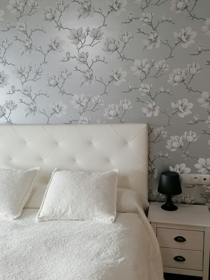 A bedroom with a silver-coloured wallpaper with white flowers behind the bed, reflecting the light in a shimmering way